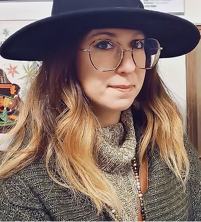 A woman wearing a black wide-brim hat, round metal-framed eyeglasses, a chunky knit beige scarf, and a textured dark green sweater, captured in a close-up portrait.